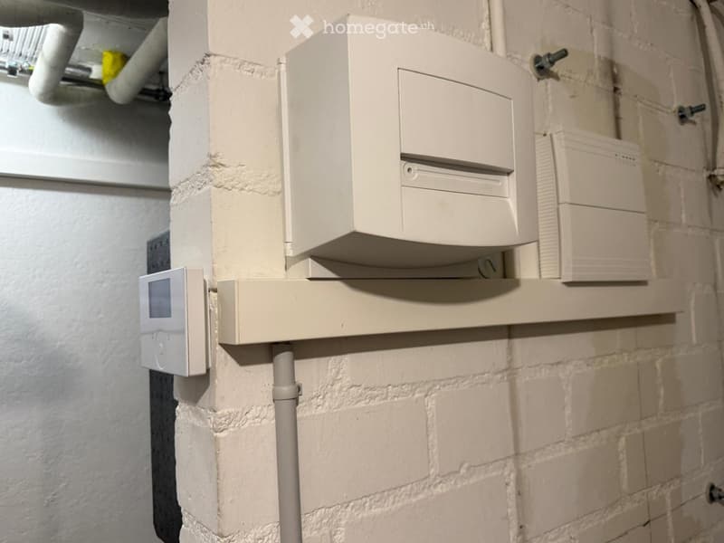 A narrow hallway with exposed brick walls, featuring a white electrical panel and a digital thermostat mounted on the wall.
