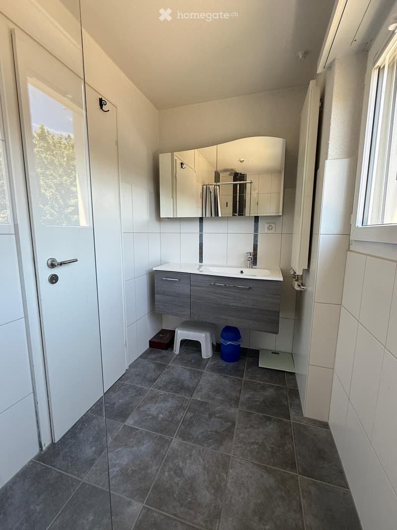 Modern bathroom with white tiled walls, grey cabinet, sink, mirror, small stool, and a blue trash bin.