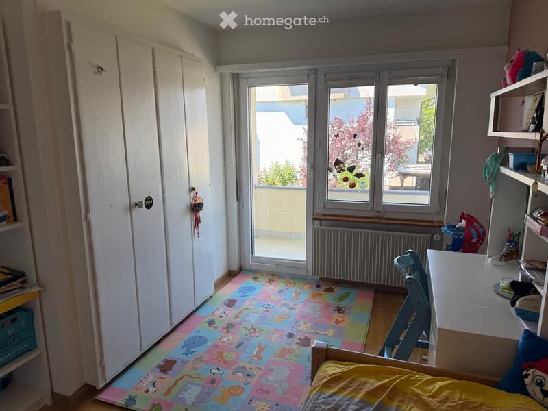 Bright bedroom with a colorful animal-themed rug, a bed with yellow bedding, a desk with a chair, shelves, and a window with a view of a tree.