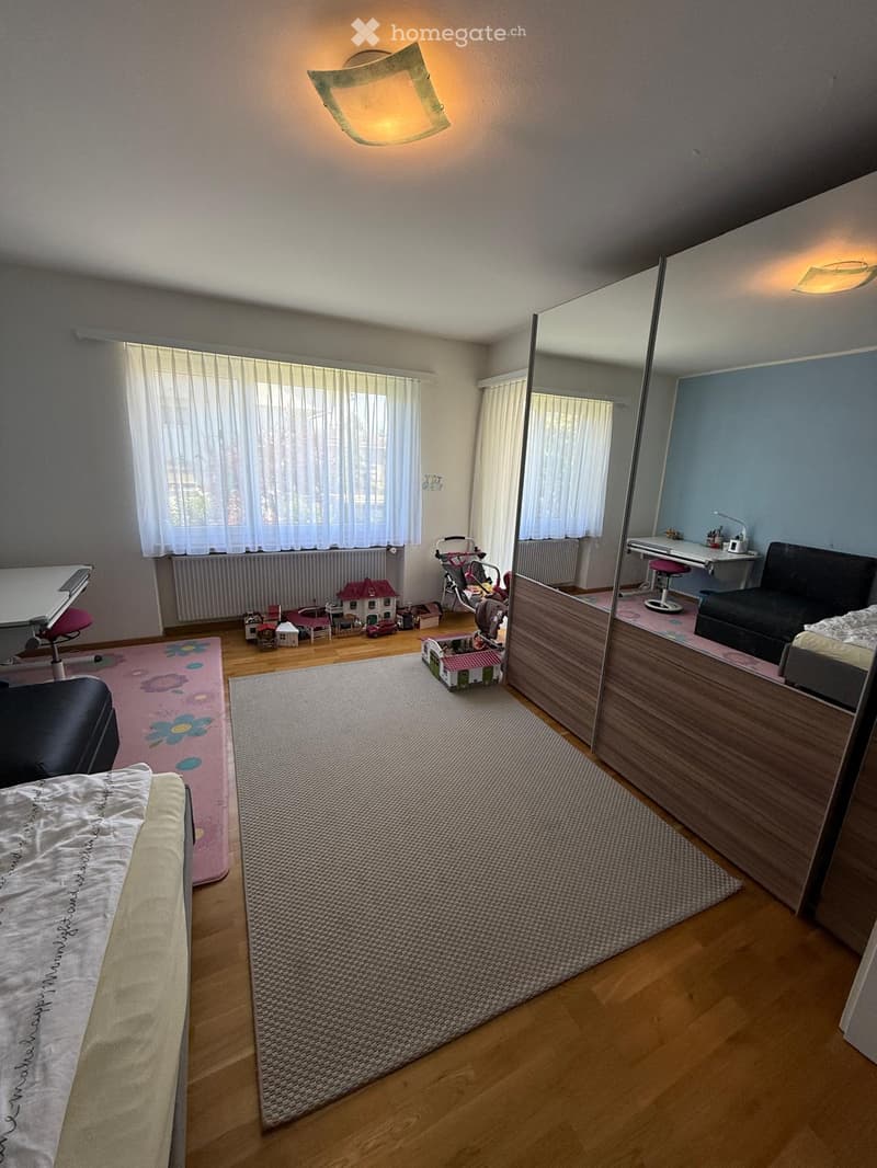 A multi-functional room with a bed, sofa, and a small desk area. The room features wooden flooring, a large mirror, and several toys scattered around.