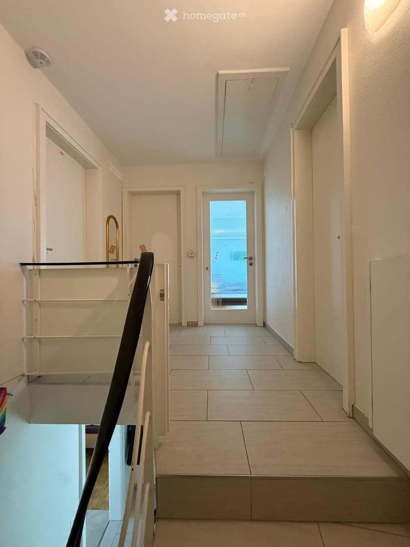 A clean, well-lit hallway with tiled flooring, white walls, and a staircase leading upwards. Features include a glass door at the end, a black handrail, and a small shelf on the left side.