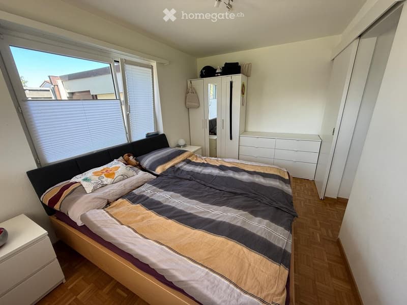 Cozy bedroom with a double bed, nightstand, wardrobe, and drawer unit. The room features wooden flooring and large windows with blinds.
