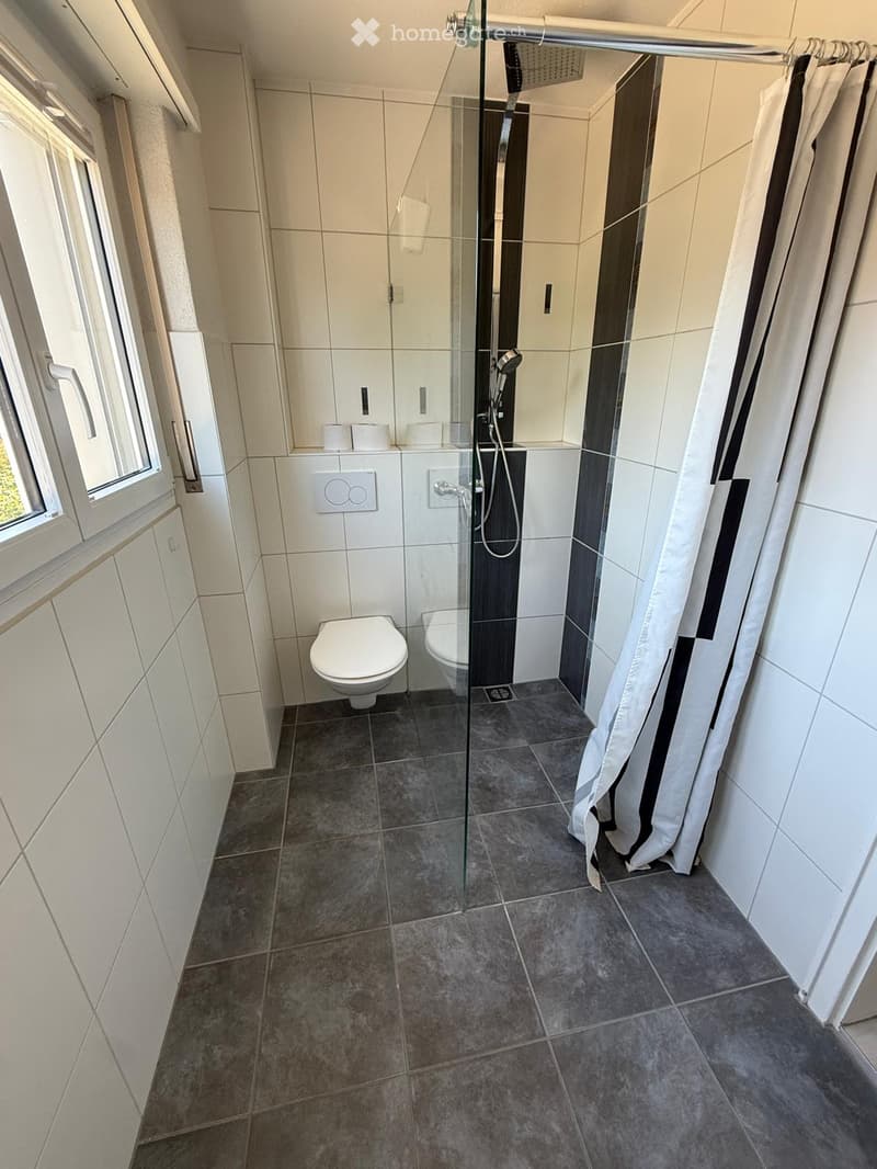 Modern bathroom with white tiles, a glass-enclosed shower, toilet, and a window with a white frame.