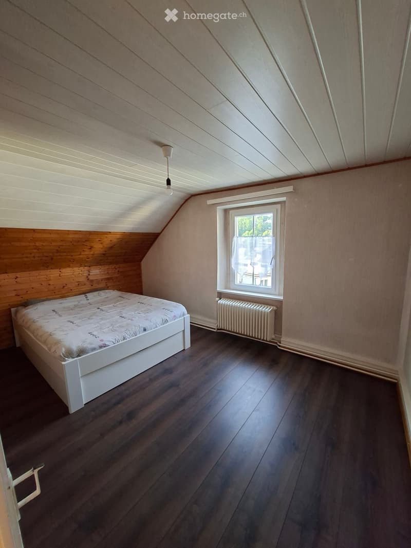 Bedroom with wooden floor, slanted ceiling, white bed with patterned bedding, radiator, window with white frame and curtains, and a hanging light fixture.