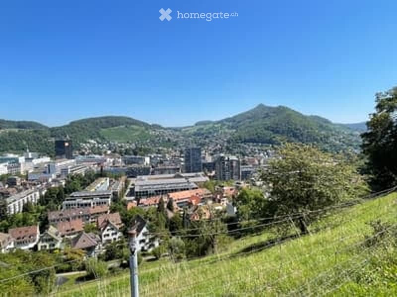 Panoramic view of a cityscape with buildings, hills, and green fields under a clear blue sky.