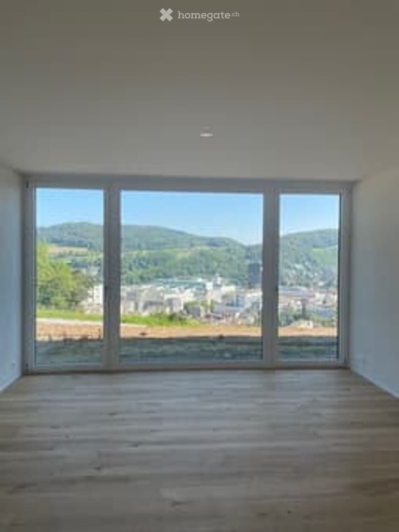 Completely empty room with large windows offering a scenic view of hills and a town. Wooden floor, white walls, and no furniture.