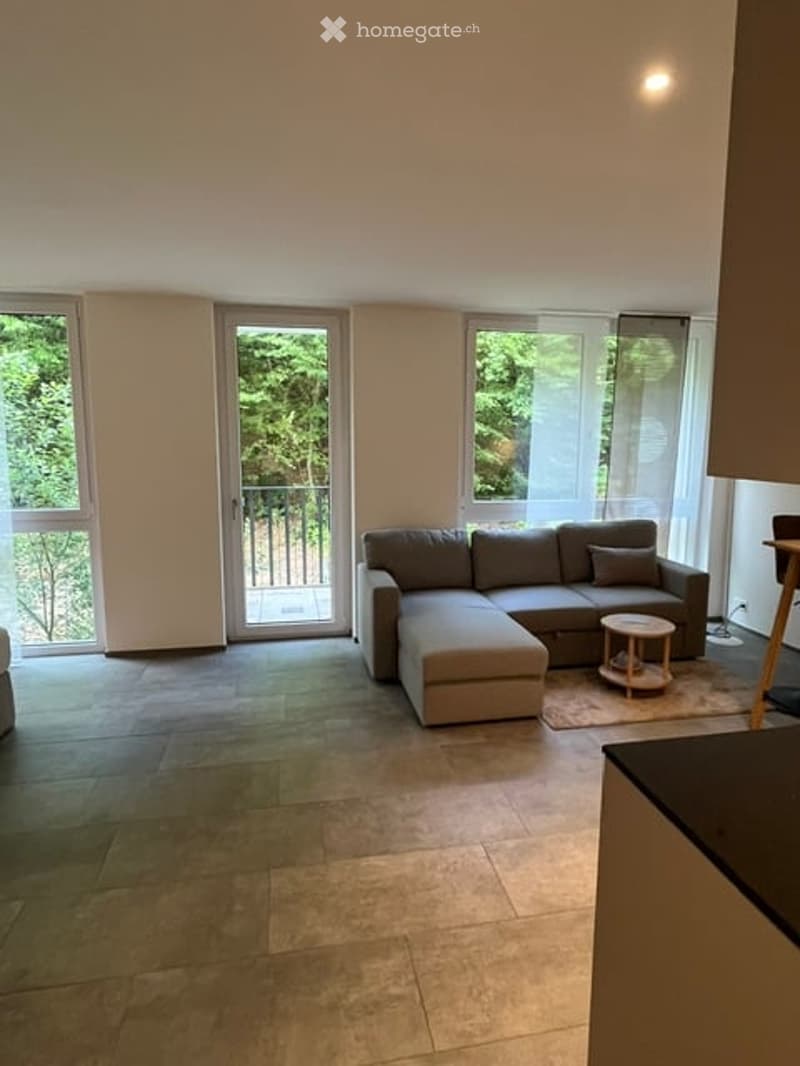 Modern living room with large windows and sliding doors leading to a balcony. Features a grey sectional sofa, a small round coffee table, and a wooden stool.
