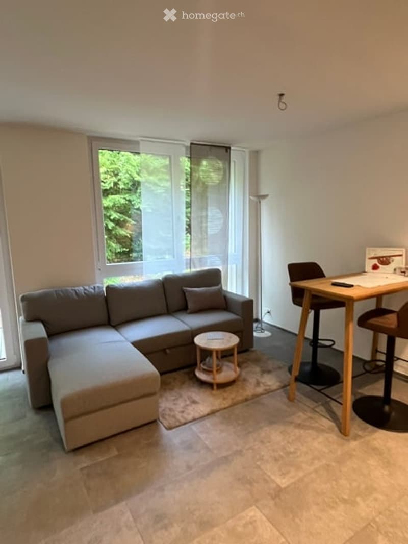 Modern living room with a grey sectional sofa, coffee table, small round table, and a dining area with a table and chairs. Large windows with blinds let in natural light.