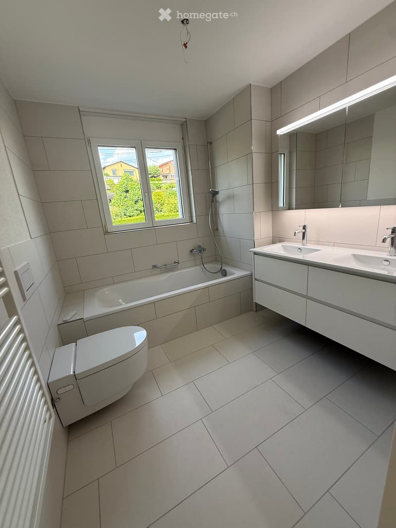 Modern bathroom with a bathtub, toilet, double sink, and large mirror. Light gray tiles and a window provide natural light.