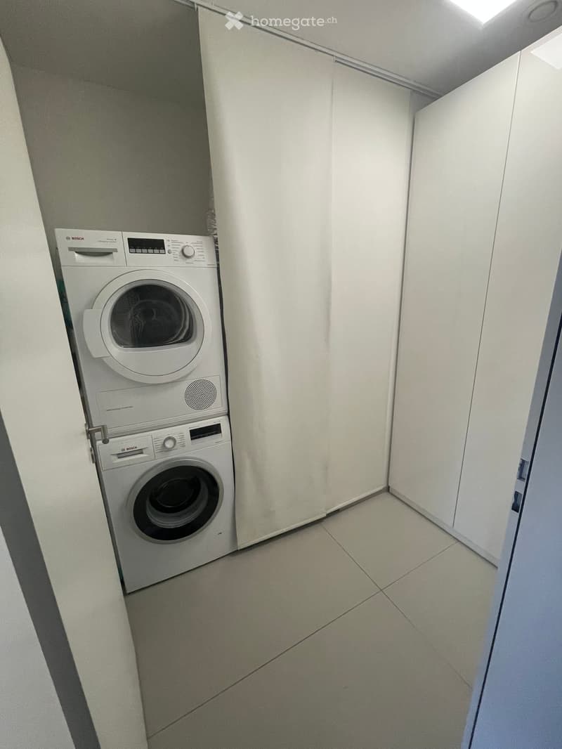 Modern bathroom with a stacked washing machine and dryer unit, white tiled floor, and white walls.