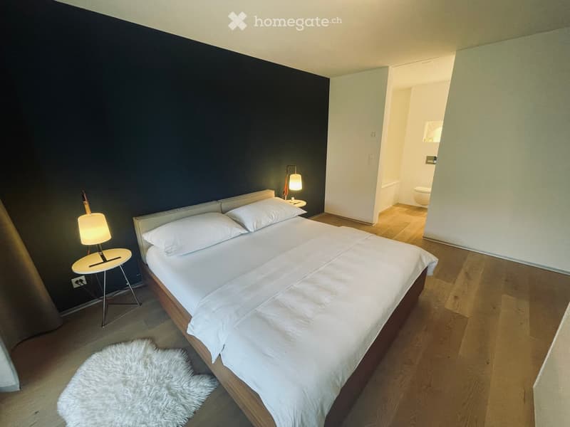 Modern bedroom with a double bed, white bedding, nightstand with lamp, small table, and a fluffy white rug on the floor. The room features dark walls and light wooden flooring, with an adjacent bathroom visible in the background.