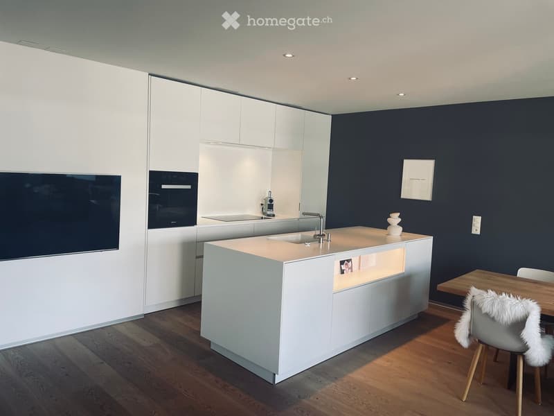 Modern kitchen with white cabinetry, dark blue accent wall, central island with built-in lighting, sink, and a small dining area with a wooden table and a white fur chair.
