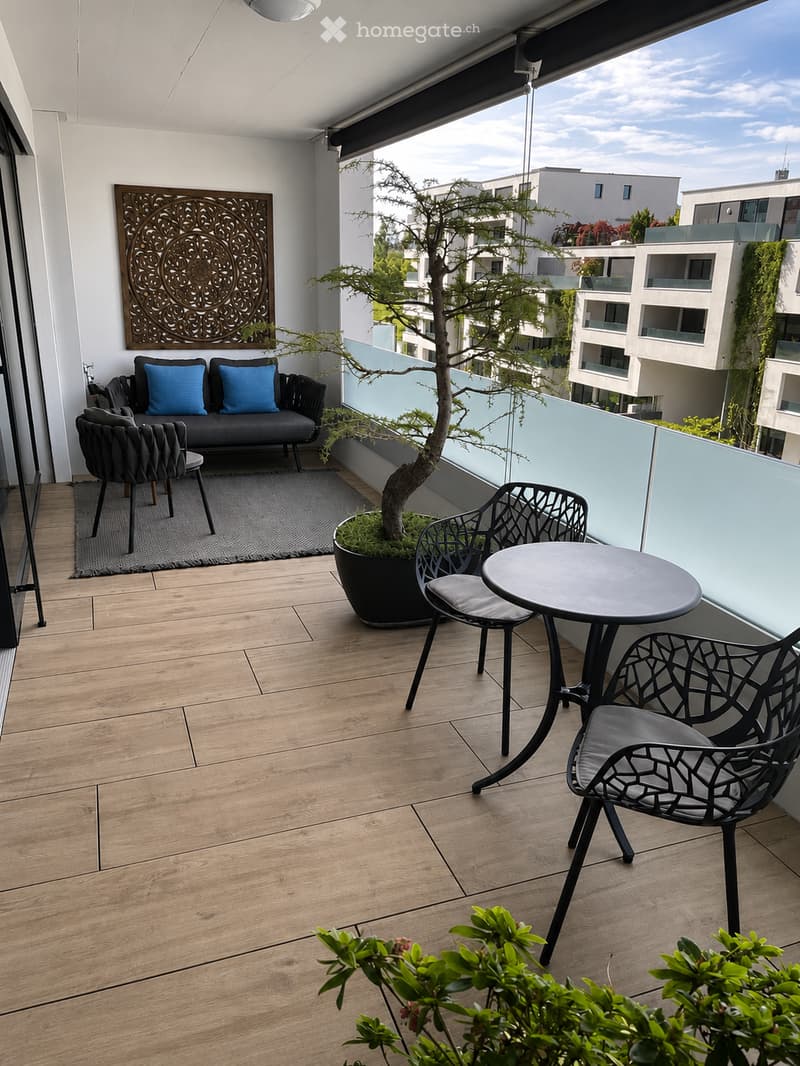 Modern balcony with wooden flooring, grey sofa with blue cushions, black metal chairs, small round table, and a potted tree.
