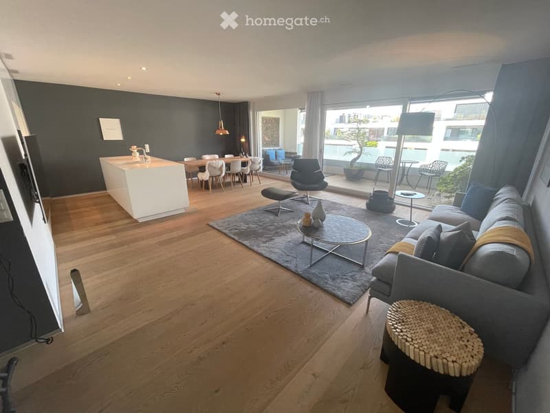 Modern living room with hardwood floors, grey sofa, coffee table, and dining area with chairs and table. Large windows with a view of the balcony and outdoor seating area.