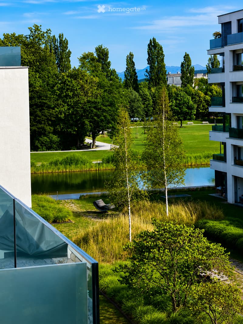 View from a balcony overlooking a landscaped garden area with a pond, trees, and modern buildings in the background.
