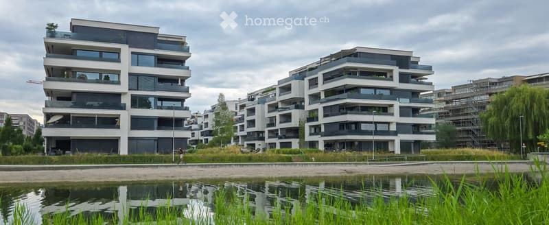 Modern multi-story residential building with balconies and large windows, situated near a water body with greenery in the foreground.