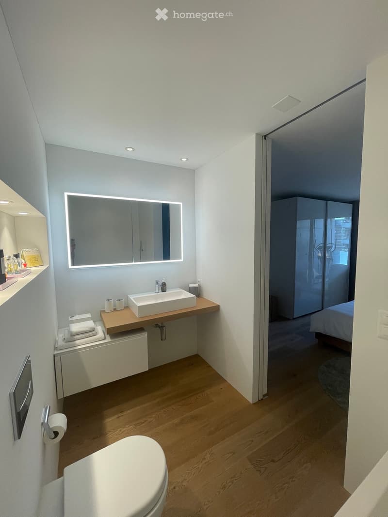 Modern bathroom with a floating wooden sink, large illuminated mirror, toilet, and a small shelf with towels and cups. The room features light wood flooring and white walls.