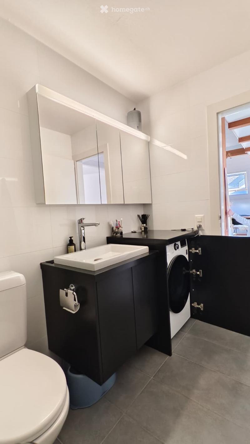 Modern bathroom with white walls, dark grey tiles, a white sink with a chrome faucet, a toilet, a large mirror with cabinets above, and a washing machine in the corner.