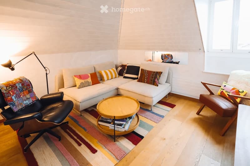 Modern living room with white walls, wooden flooring, a sectional sofa, a coffee table with books, a leather armchair, and a floor lamp.