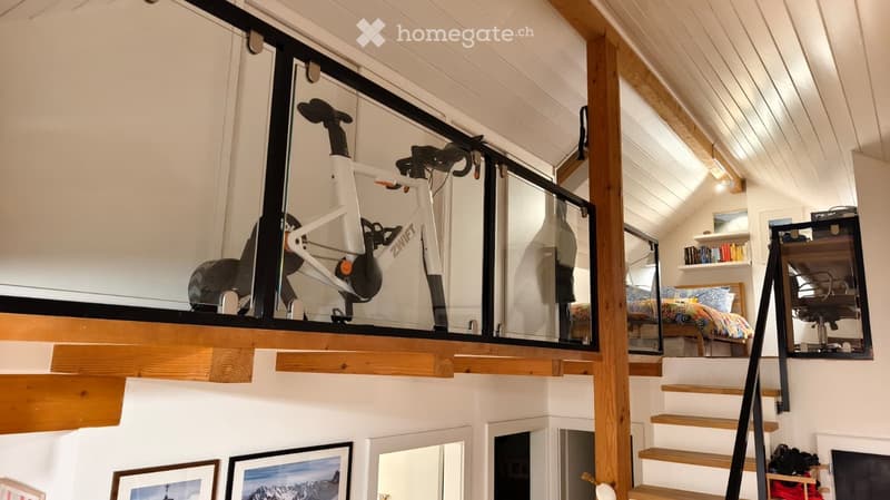 Loft-style room with a raised platform holding a bicycle, a chair, and a bed with colorful bedding. Wooden stairs lead up to the platform, and shelves and picture frames decorate the walls.