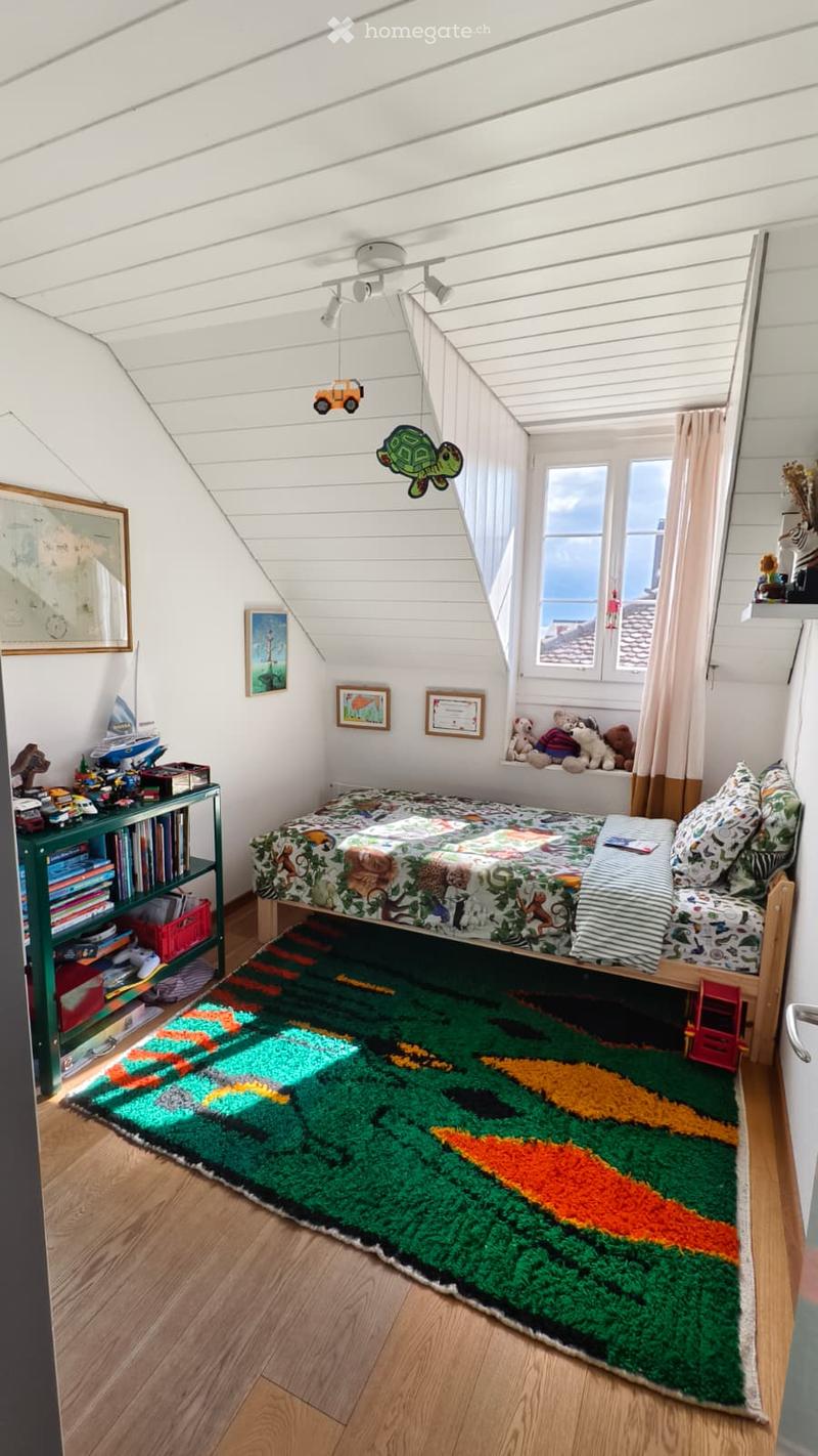 Cozy children's bedroom with a wooden bed, colorful quilt, green bookshelf, and a vibrant rug. The room features slanted ceilings, large windows with curtains, and various toys and decorations.