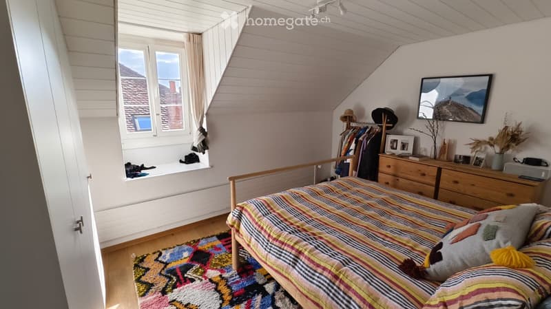 Cozy attic bedroom with wooden bed, colorful striped bedding, patterned rug, dresser, mirror, and decorative items on the windowsill.