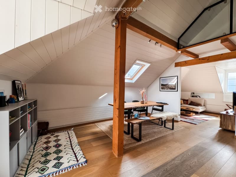 Bright, open loft-style living space with wooden floors, exposed wooden beams, skylight, and large windows. Features a sofa, coffee table, dining table with chairs, bench, shelf, and decorative items.