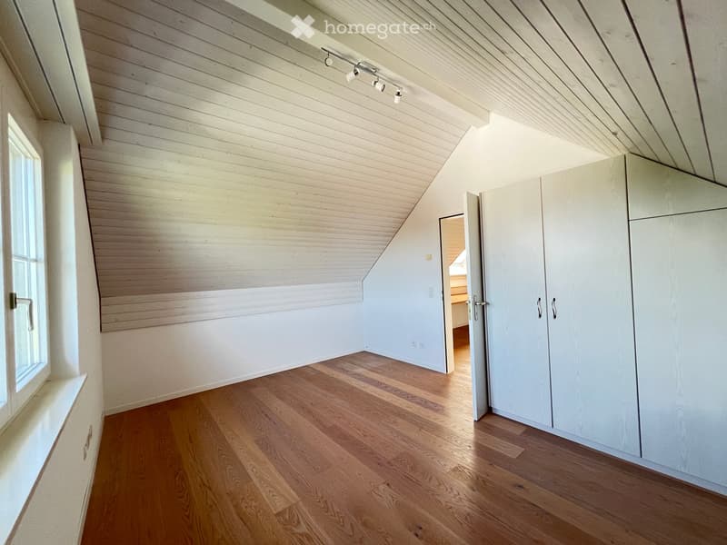 An empty attic room with wooden floors, white walls, and built-in storage cabinets. The room features exposed wooden beams on the ceiling and a large window on one side.