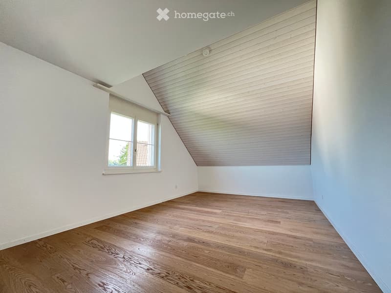 Empty room with wooden flooring, white walls, and a slanted ceiling. Features a single window with white frame and blinds.