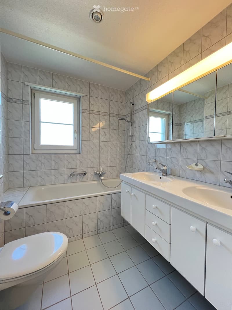 Modern bathroom with marble tiles, a bathtub, toilet, double sink, and a large mirror with built-in lighting. Features a window for natural light and a sleek, contemporary design.