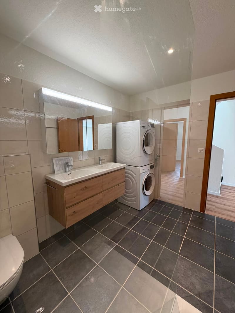 Modern bathroom with dark tiled floor, double sink vanity with wooden cabinet, large mirror, and stacked washer/dryer unit.
