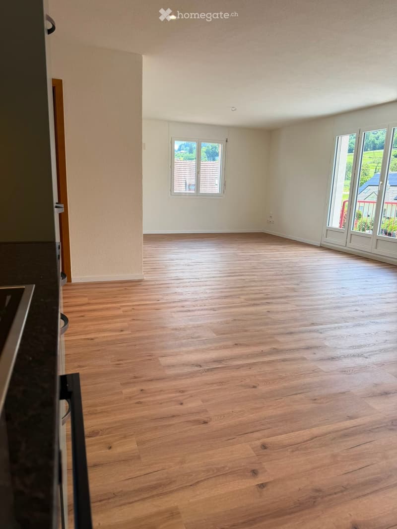 Empty room with wooden flooring, white walls, and large windows, including a sliding glass door leading outside.