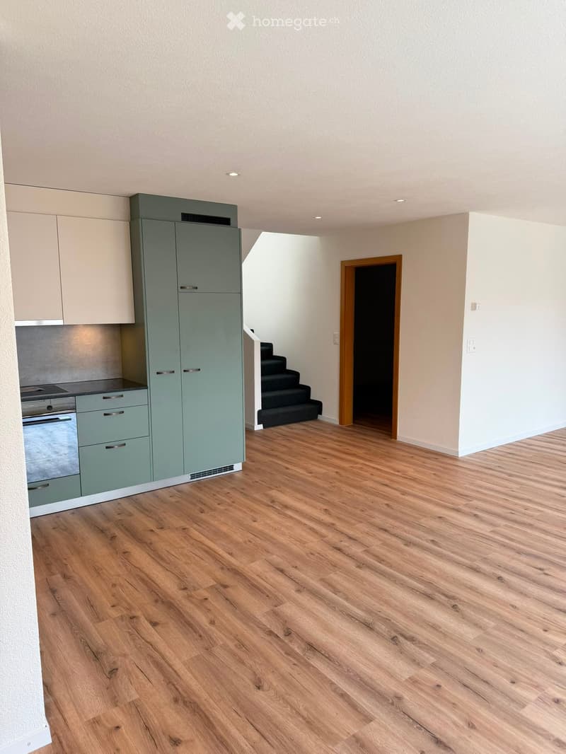 Empty room with light wooden flooring, white walls, and a small kitchen area with cabinets and appliances visible in the background.