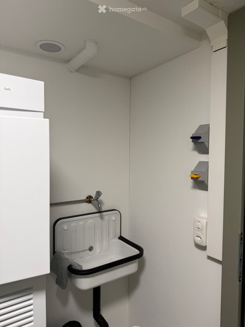 Compact bathroom with a white sink, chrome faucet, and a grey towel hanging on the side. The walls are light grey, and there are electrical outlets and a small shelf with items on the right side.