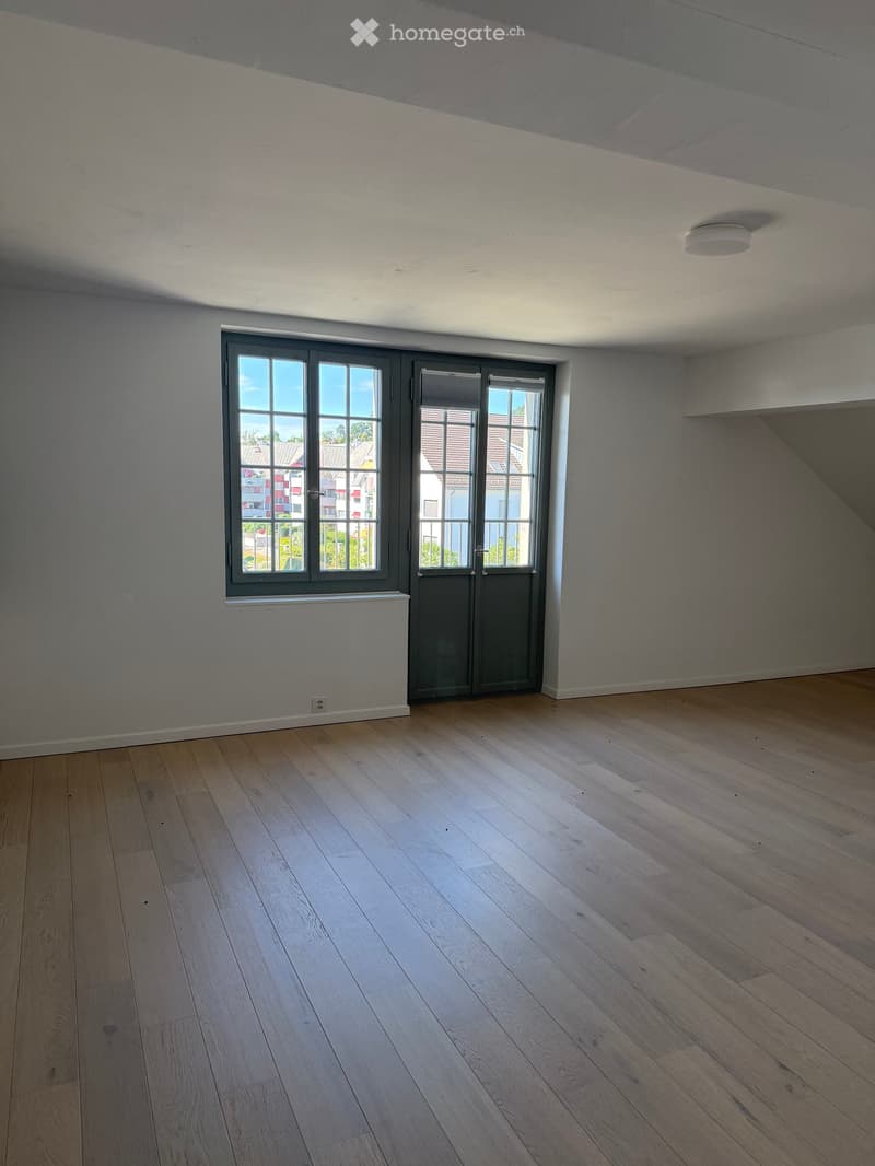 Empty room with light wooden flooring, grey walls, and large windows with grey frames, allowing natural light to enter.