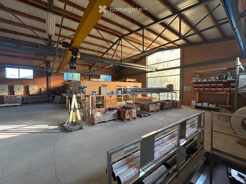 Empty warehouse space with exposed wooden beams, brick walls, large windows, and scattered storage items and materials.