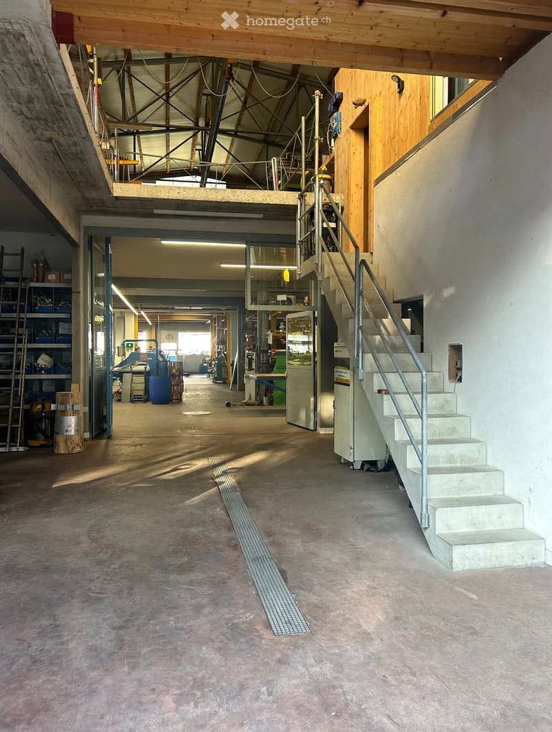 Empty industrial-style room with concrete floors, exposed wooden beams, and a staircase leading to an upper level. The space includes a large open area with scattered storage items and equipment in the background.