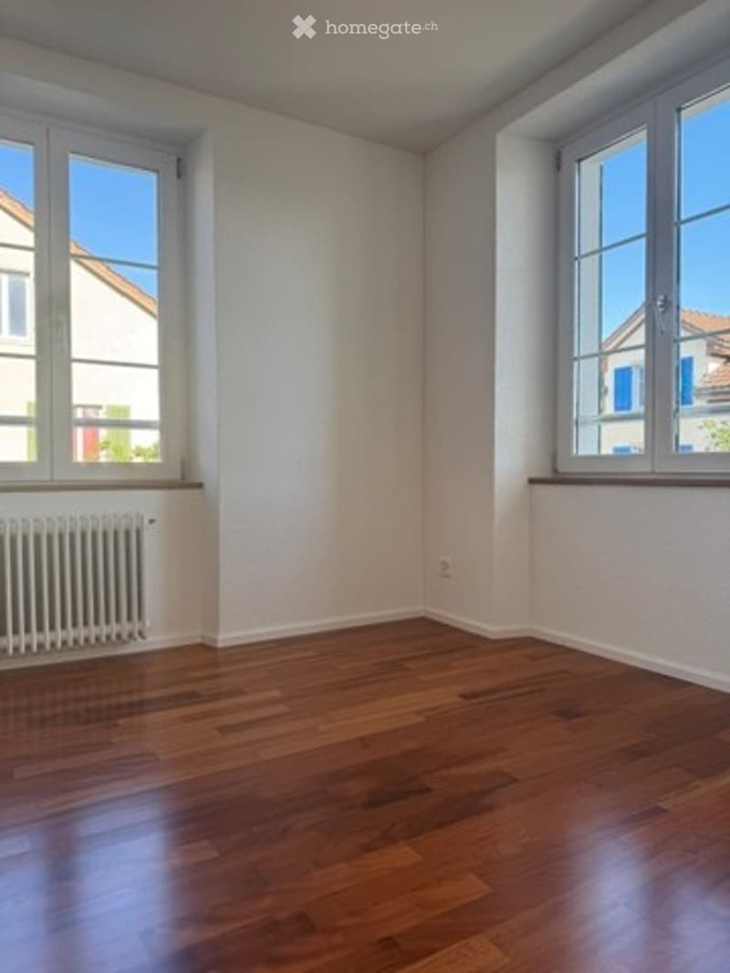 Empty room with wooden flooring, white walls, and two windows with white frames. A radiator is visible under one window.