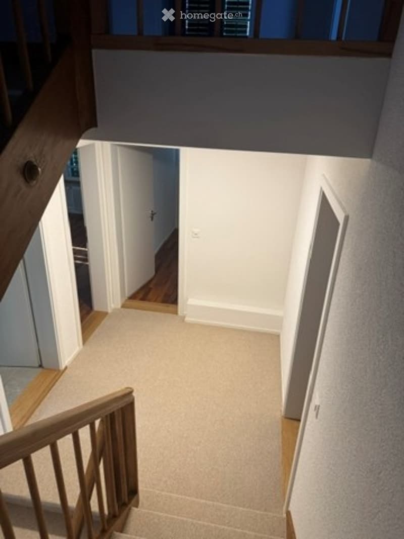 Spacious, well-lit hallway with carpeted stairs leading upwards, white walls, wooden railings, and a visible doorway at the end.