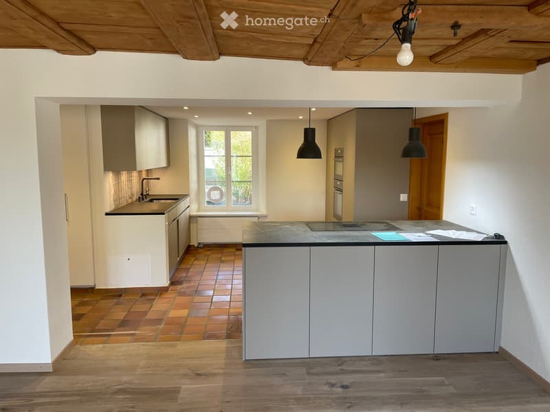 Modern kitchen with light grey cabinets, a central island, and a tiled backsplash. Features pendant lights, a sink, and an oven.