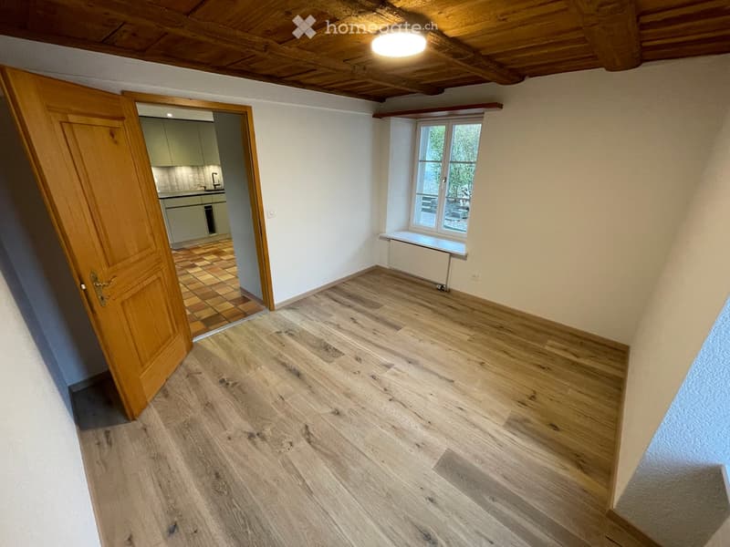 Empty room with wooden flooring, white walls, and a wooden door leading to a kitchen area. The room has a window with a radiator below it and a visible light fixture on the ceiling.