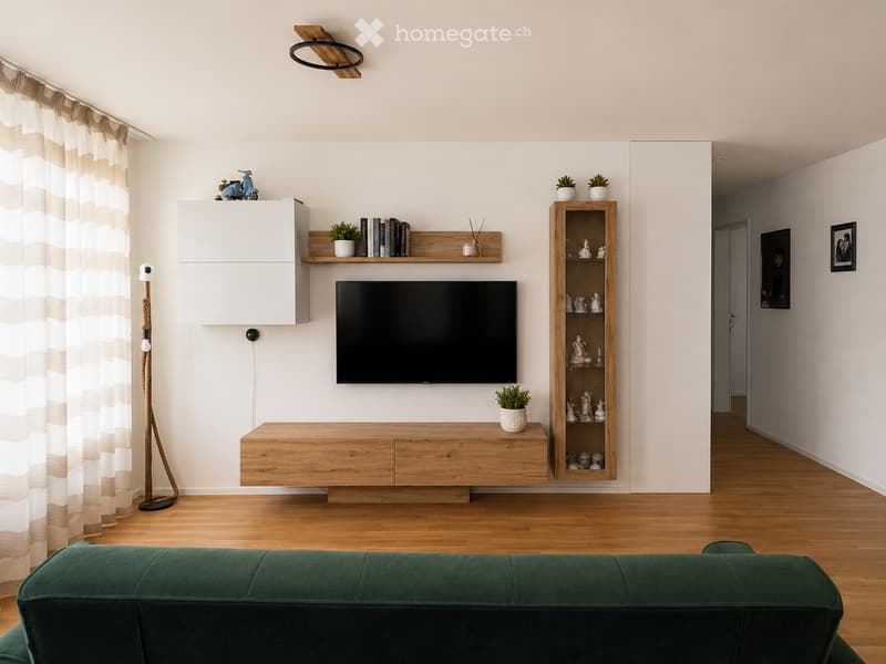 Modern living room with a green sofa, wooden cabinet, shelves with books and potted plants, a television mounted on the wall, and a floor lamp with a rope design.
