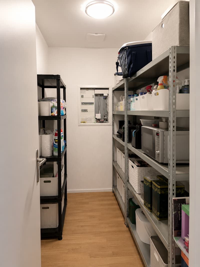 A well-organized storage room with wooden flooring, white walls, and multiple shelves filled with cleaning supplies, a microwave, and storage bins.