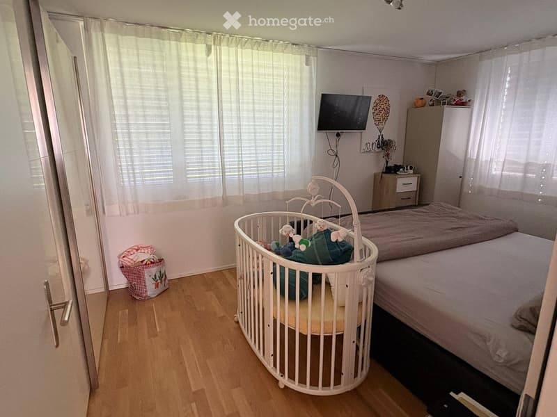 A well-lit bedroom with a double bed, a crib, a nightstand, and a television mounted on the wall. The room features wooden flooring and large windows with curtains.
