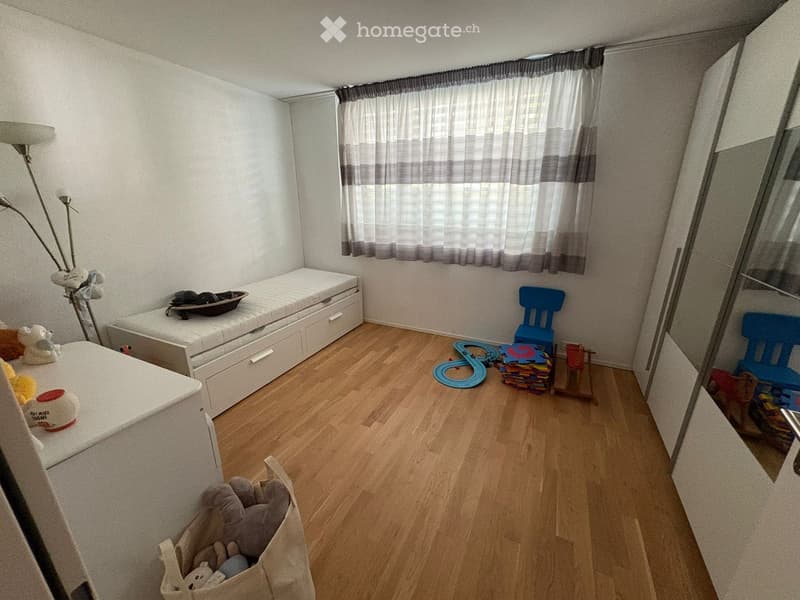 A small, well-lit bedroom with wooden flooring, a single bed with a mattress, a chair, toys, and a mirror. The room features a window with curtains and a lamp near the bed.