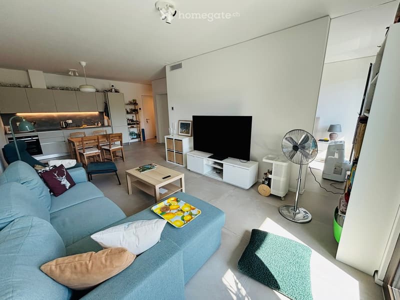 A modern, open-plan living and dining area with a blue sofa, coffee table, dining table with chairs, kitchen area, and a television. The space is well-lit with natural light and features contemporary decor.