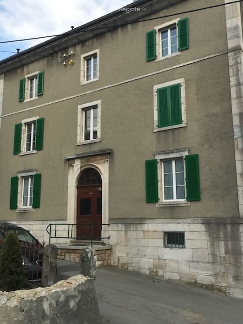 A three-story residential building with beige walls, green shutters, and white-framed windows. The entrance features a wooden double door with a small staircase leading up to it. A parked car is visible in the foreground.
