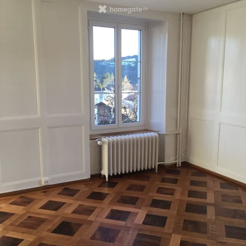 Empty room with wooden parquet flooring, white walls, a window with a view, and a radiator below the window.