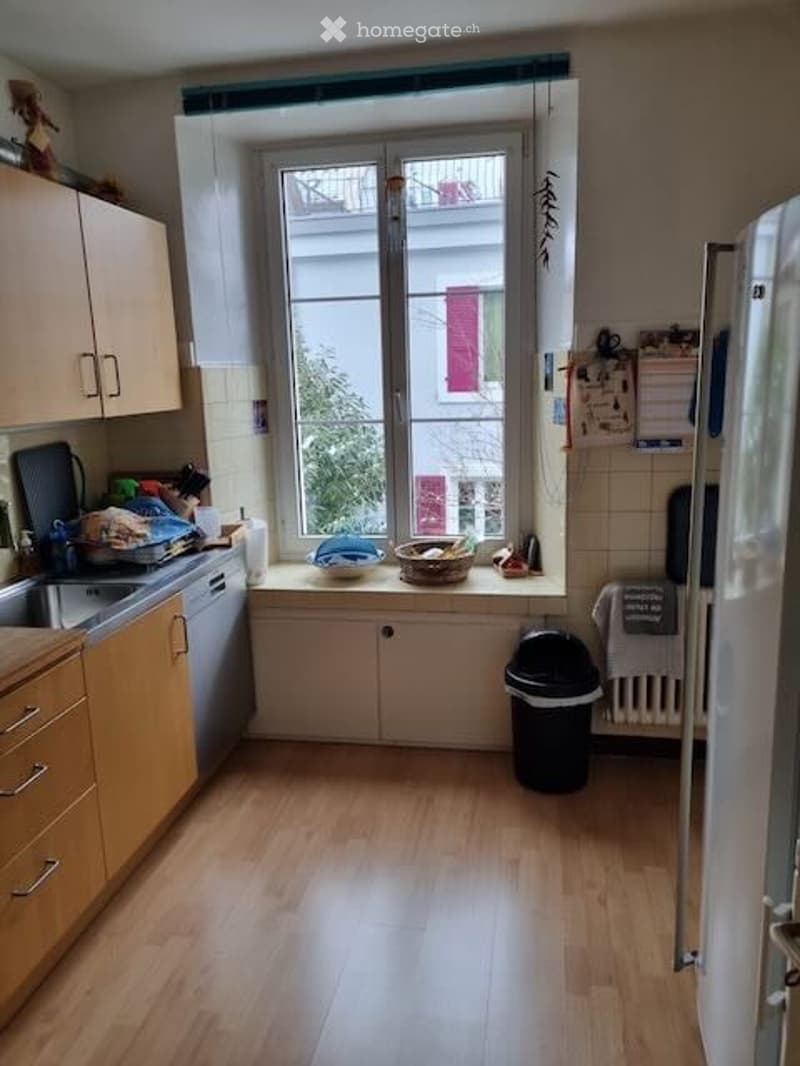 Bright kitchen with light wooden cabinets, a window above the sink, and a dishwasher. The floor is light wood, and there is a trash bin and a basket on the windowsill.