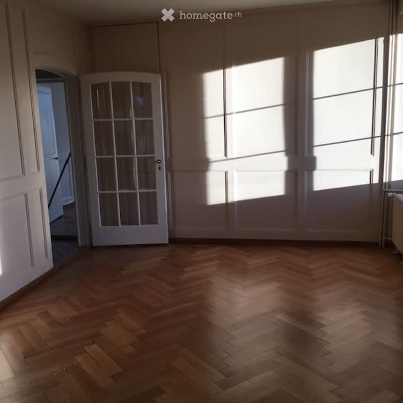 Empty room with wooden herringbone flooring, white walls, large windows, and a glass door leading to another space.
